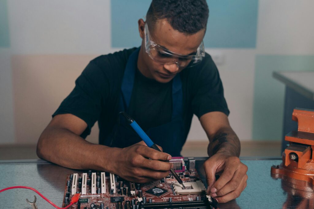 A technician wearing safety glasses works intently on a computer motherboard with a soldering iron.